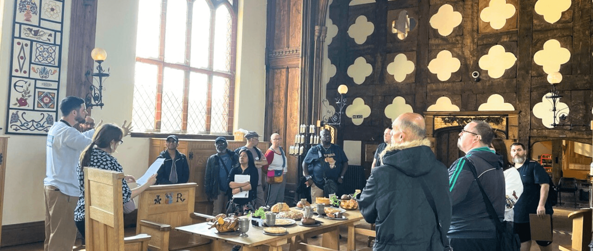 Manchester Deaf Centre – A group of people are gathered around a banqueting table in a great Tudor Hall. A man is on the left of the photo is signing.