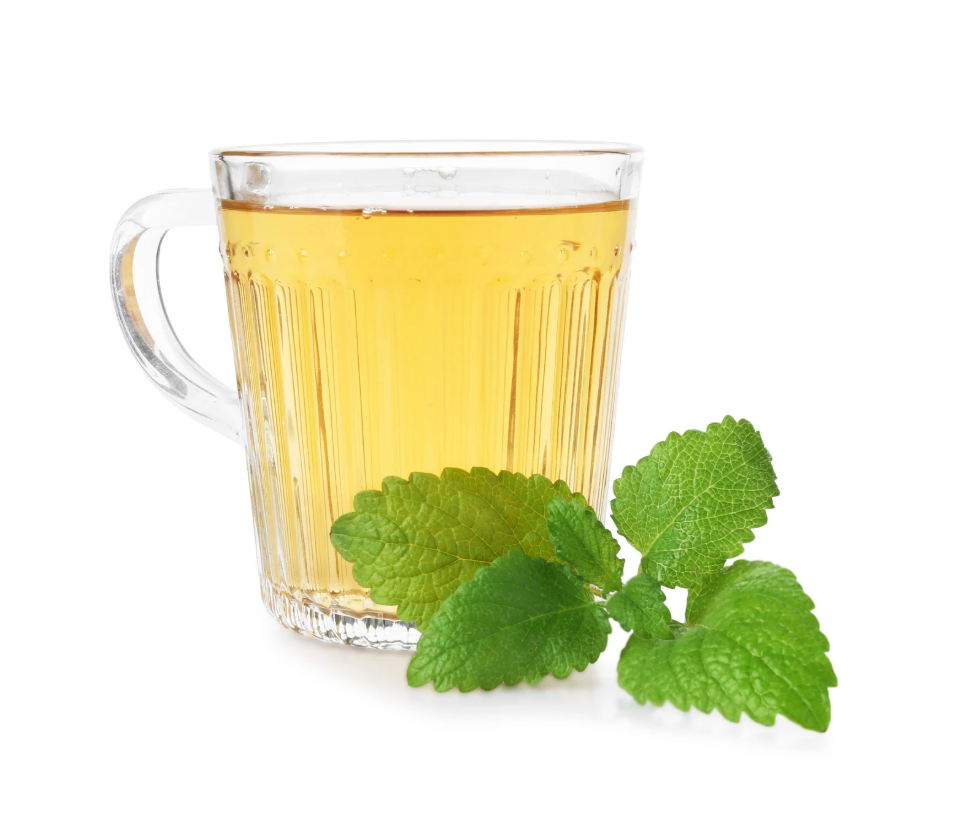 A glass of pale yellow liquid next to some lemon balm leaves